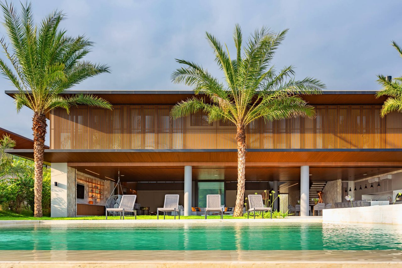 Fachada contemporânea em brises de alumínio amadeirado e superfícies envidraçadas, com palmeiras e piscina diante do mar